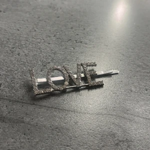Glittrig hårspänne med LOVE-text - Snyggt hårspänne i silverfärg med ordet LOVE format av glittrande stenar. Perfekt accessoar för att ge din frisyr extra bling och personlighet. Spännet är smalt och stilrent, med tydliga bokstäver som fångar ljuset.
