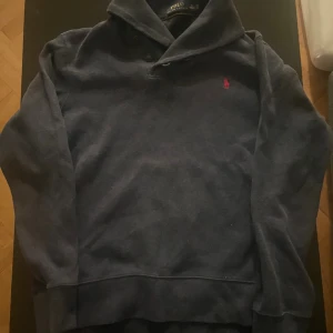 Mörkblå tröja från Polo Ralph Lauren - Snygg mörkblå tröja från Polo Ralph Lauren i storlek S. Tröjan har lång ärm, klassisk krage med knappar och den ikoniska röda logon broderad på bröstet. Perfekt för dig som gillar stilrena och tidlösa plagg.