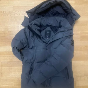 Mörkgrå dunjacka från Canada Goose - Säljer en mörkgrå dunjacka från Canada Goose med stor huva och snyggt svart patch på ärmen. Jackan har dragkedja och knappar framtill, samt två praktiska fickor. Perfekt för kalla dagar och har en riktigt schysst oversized look. En knapp saknas som inte är visad