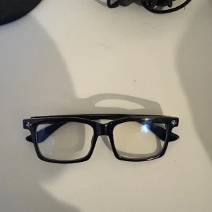 Svarta glasögon med silverdetaljer - Snygga svarta glasögon från Chrome Hearts med rektangulära bågar och coola silverdetaljer på skalmarna. Bågarna har unika mönster och små stjärnor i metall, vilket ger en lyxig och edgy vibe. Perfekta för dig som vill sticka ut med din stil.