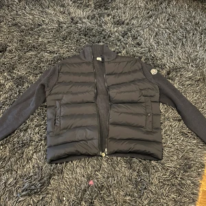 Svart pufferjacka från Moncler - Svart pufferjacka från Moncler med stickade ärmar och quiltad kropp. Jackan har dragkedja framtill, två fickor med knappstängning och Moncler-logga på vänster ärm. Materialmix av polyester och stickad ull ger en schysst kontrast. INGA SPRÅ AV ANVÄNDNIN! Jackan är i nyskick