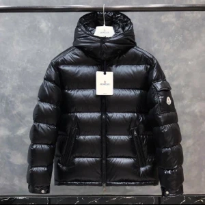 Svart Moncler pufferjacka med huva - Svart pufferjacka från Moncler med glansig finish, huva och två stora fickor med dragkedja. Jackan har Moncler-logga på ärmen och coola detaljer som quiltad design och innerfoder med serietidningsmotiv. Perfekt för dig som vill ha en snygg och varm jacka.