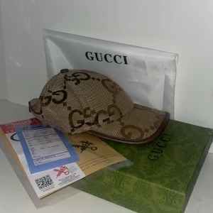 Gucci beige keps med GG-mönster - Snygg beige keps från Gucci med klassiskt GG-mönster i brunt. Kepsen har justerbar rem i brunt skinn baktill och är tillverkad i bomull och polyester. Perfekt för dig som vill ha en lyxig och trendig accessoar med tydlig designerstil.