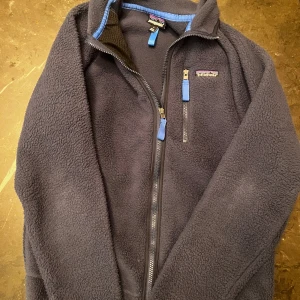 Mörkblå fleecejacka från Patagonia - Mysig mörkblå fleecejacka från Patagonia med hel dragkedja och bröstficka med blå zip-pullers. Jackan har lång ärm och stårkrage, perfekt för kalla dagar. Snygga detaljer i blått vid kragen och fickan. Klassisk outdoor-look och riktigt skön passform.