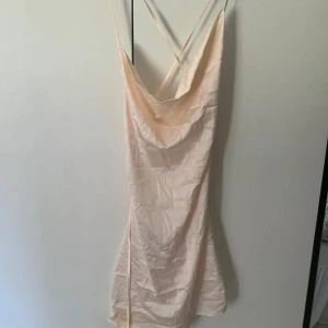 Ljusbeige kort klänning från Parisian - Supersöt ljusbeige kort klänning från Parisian med tunna axelband som korsas i ryggen och snygg draperad urringning. Klänningen har en figurnära passform och är tillverkad i ett mjukt, glansigt material med stretch. Perfekt för dig som gillar minimalistisk och trendig stil.