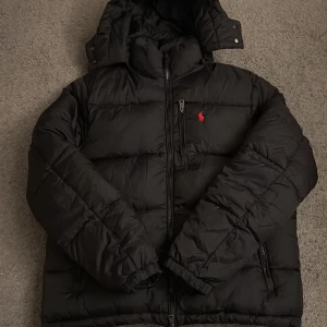 Svart pufferjacka från Polo Ralph Lauren - Svart pufferjacka från Polo Ralph Lauren med röd broderad logga på bröstet. Jackan har huva, flera fickor med dragkedja och elastiska muddar. Materialet är syntet och dun, perfekt för kalla dagar. Klassisk och stilren design med Polo-detaljer på dragkedjan.