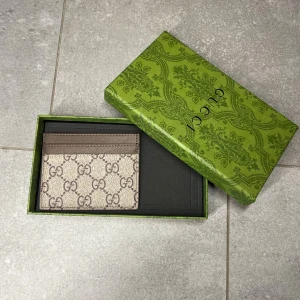  beige plånbok/korthållare - Snygg beige plånbok med klassiskt mönster i brunt. Plånboken har flera fack för kort och levereras i original grön ask med dekorativt mönster. Perfekt för dig som gillar stilrena accessoarer med lyxig känsla.