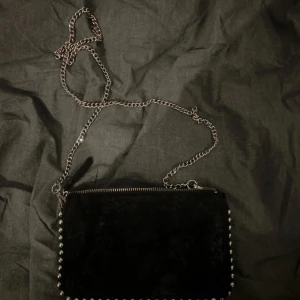 Svart mocka axelväska med kedja - Snygg svart axelväska i mocka med silverfärgad kedja som axelrem och coola nitar längs kanterna. Väskan har tre fack med dragkedja och en stilren, rektangulär form. Perfekt för att bära det viktigaste och ge outfiten en edgy touch.