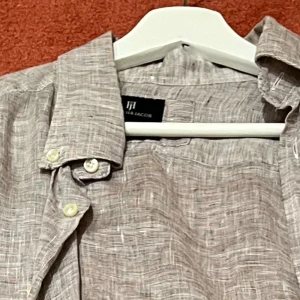 Grå linneskjorta från H & Jacob - Snygg grå skjorta i linne från H & Jacob med diskret rutigt mönster. Skjortan har klassisk krage, långa ärmar och knappar framtill samt vid manschetterna. Perfekt för dig som gillar chill och stilren look.