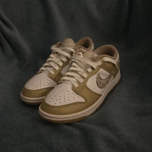 Nike Dunk Low beige och vit paisley - Nike Dunk Low sneakers i beige och vitt gula med paisleymönstrad swoosh. Skorna har snörning, perforerad tå och platt sula. Ovandelen är i skinn och syntet, med en låg siluett som ger en clean och stilren vibe. Perfekt för dig som gillar unika detaljer och streetwear.