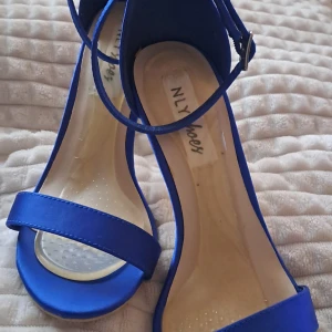 Blå klackade sandaler från NLY Shoes - Snygga blå sandaler med hög klack från NLY Shoes. Skorna har öppen tå, smal rem över foten och justerbart spänne vid ankeln. Materialet har en satinliknande finish som ger en lyxig känsla. Perfekt för dig som vill sticka ut med färg och stil.
