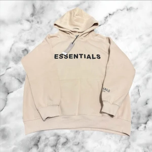 Beige Essentials Fear of God hoodie - Snygg beige hoodie från Essentials Fear of God i storlek M. Tröjan har stor svart logga på bröstet och mindre tryck på ärmen. Klassisk huva och känguruficka framtill. Mjuk bomullsmix som är skön att bära och passar perfekt till streetwear-stilen.