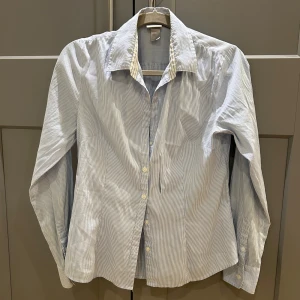 Blårandig skjorta från H&M, strl 40 - Snygg skjorta från H&M i storlek 40 med tunna blå och vita ränder. Klassisk krage, långa ärmar och knäppning framtill med vita knappar. Kvinnlig passform 
