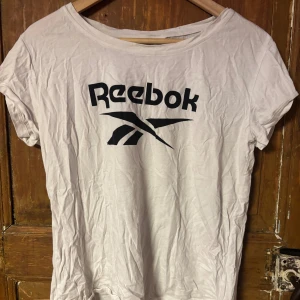 Vit Reebok t-shirt med logga - Vit t-shirt från Reebok med svart tryckt logga på bröstet. Klassisk rund hals och korta ärmar. Mjuk och lätt bomullskänsla, perfekt för en sportig look. Enkel design utan mönster eller extra detaljer.