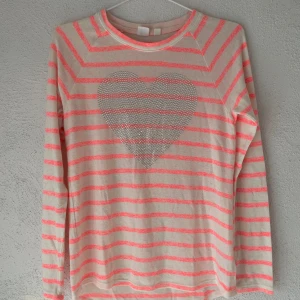 Randig tröja med hjärta i strass - Beige tröja med neonrosa ränder och ett stort hjärta i strass framtill. Långärmad och i mjukt bomullsmaterial, perfekt för dig som gillar färg och detaljer. Snygg till jeans eller kjol för en chill look.