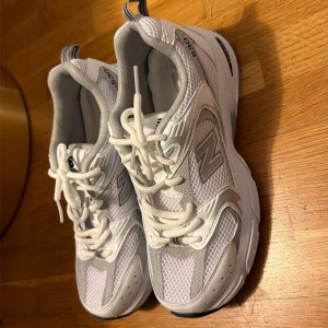 New Balance 530 vita sneakers - Säljer ett par New Balance 530 sneakers i vitt och silver med mesh och syntetdetaljer. Skorna har en chunky sula, svarta detaljer undertill och klassisk snörning. Perfekta för dig som gillar sportig och trendig stil.