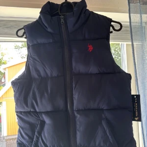 Mörkblå dunväst från U.S. Polo Assn XS - Snygg mörkblå dunväst från U.S. Polo Assn i storlek XS. Västen har hög krage, dragkedja framtill och broderad röd logga på bröstet. Perfekt för lager-på-lager och håller dig varm med sin quiltade design.