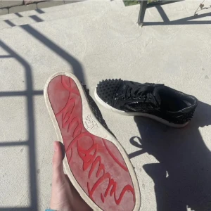 Christian Louboutin svarta sneakers med nitar - Säljer ett par svarta sneakers från Christian Louboutin med ikonisk röd sula och coola svarta nitar över hela skon. Skorna har snörning, rund tå och en platt vit sula. Materialet är en mix av skinn och mocka med glansiga detaljer. De är i storlek 39 och är använda få tal gånger  fattas två spikar men de är enkelt att fixa  box och kvitto tillkommer  köptes för 2 år sen i purto banos 