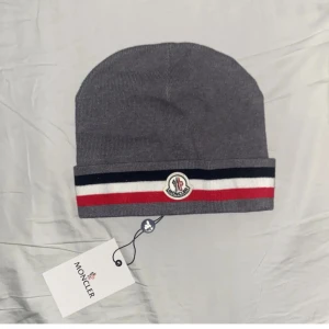 Grå Moncler mössa med ränder - Snygg grå mössa från Moncler med klassiska ränder i svart, vitt och rött längs kanten. Mössan har Moncler-logga framtill och är ribbstickad för en clean look. Perfekt accessoar för kyliga dagar och streetstyle.