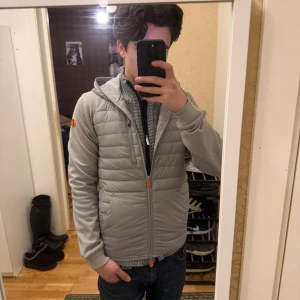 Save the Duck Cardigan - Säljer en grå cardigan från Save The Duck i storlek S. Jackan har huva, orange detaljer på dragkedjorna och ett snyggt emblem på ärmen. Materialet är mestadels polyester och nylon, med quiltad framsida och mjuka ärmar.