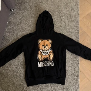 Svart Moschino hoodie med björntryck - Svart hoodie från Moschino med ikoniskt björntryck. Tröjan har huva och är tillverkad i mjuk bomull, perfekt för en chill och trendig look. Långärmad modell som passar dig som gillar streetstyle.