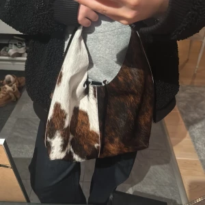 Brunvit axelväska i koskinn-look - Cool axelväska med unik koskinn-look i brunt, vitt och svart från other stories. Andvänd 1 eller 2 gånger. Materialet är pälsimitation. Nypris 1890.