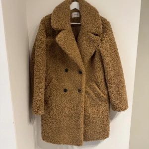 Brun teddyjacka från Reserved - New. No sign of use. Säljer en brun teddyjacka med bred krage och dubbelknäppning framtill. Jackan har två sidofickor och är helfodrad med slät insida. Den har en loose passform och är perfekt för kalla dagar. Färgen är varm brun och materialet är mjukt teddy.
