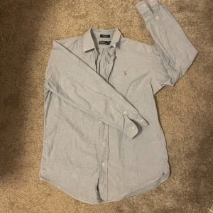 Ljusblå skjorta från Polo Ralph Lauren - Snygg ljusblå skjorta från Polo Ralph Lauren med klassisk krage och broderad logga på bröstet. Skjortan har långa ärmar, vita knappar och är tillverkad i mjuk bomull. Perfekt för dig som gillar stilrena och tidlösa plagg. Pris kan diskuteras vid snabbaffär