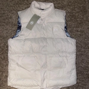 Vit dunväst från Canada Goose - Snygg vit dunväst från Canada Goose med quiltad design och hög krage. Utsidan är stilren med diskret logga på bröstet och två stora fickor framtill. Insidan har ett unikt mönster i blått och svart. Perfekt för lager-på-lager under kyliga dagar.