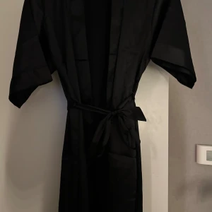 Svart satin morgonrock med knytband - Svart satin morgonrock i omlottmodell med knytband i midjan och korta ärmar. Klänningen har en stilren look och är tillverkad i ett mjukt, glansigt material som ger en lyxig känsla. Perfekt för dig som gillar minimalistisk stil och vill ha något enkelt men snyggt.  Från missäy