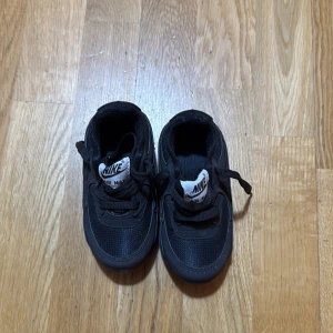 Svarta Nike Air Max sneakers strl 23 - Säljer ett par svarta Nike Air Max sneakers med klassisk synlig luftkudde i sulan. Skorna har snörning, rund tå och är tillverkade i syntetmaterial med meshdetaljer för extra ventilation. Snyggt Nike-märke på plösen och sidan. Perfekt för dig som gillar sportig stil.
