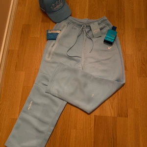 Nike NOCTA ljusblå mjukisbyxor M - Säljer ett par ljusblå mjukisbyxor från Nike NOCTA i storlek M. Byxorna har snörning i midjan, dragkedjeficka på sidan och NOCTA-logga samt Nike Swoosh framtill. Materialet är mjukt och skönt, perfekt för chill eller streetwear.