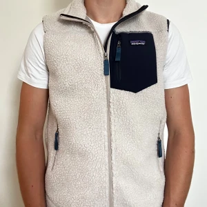 Vit fleeceväst från Patagonia M - Säljer en vit fleeceväst från Patagonia i storlek M. Västen har en mjuk och fluffig yta, hel dragkedja framtill och en mörkblå bröstficka med dragkedja. Två sidofickor med dragkedja och hög krage för extra värme. Perfekt för lager-på-lager under kyliga dagar.