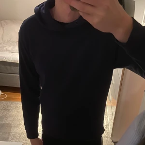 Mörkblå hoodie med huva och ficka - Säljer en stilren mörkblå hoodie med stor huva och klassisk magficka. Tröjan har långa ärmar och ribbade muddar vid ärmslut och nederkant. Perfekt för chill dagar och enkel att matcha med jeans eller joggers.