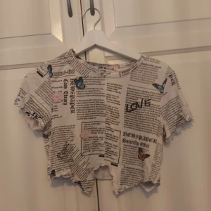 Vit croppad topp med tidningsprint FB Sister - ⚠️ Skriv till mig innan köp!⚠️Säljer en vit croppad topp från FB Sister i storlek M. Toppen har tidningsmönster med text och fjärilar i blått och brunt, samt rå kant nertill. Materialet är mjuk bomull och modellen har korta ärmar. Perfekt för dig som gillar unika prints och streetstyle.