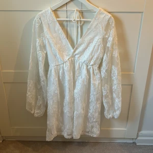 Vit blommig kortklänning Gina Tricot - Supersöt vit kortklänning från Gina Tricot i storlek XS. Klänningen har transparenta långa ärmar, v-ringad omlottfront och är täckt med broderade blommor. Den har knyt i nacken och ett flowigt fall som ger en romantisk vibe. Perfekt för student eller sommarens alla tillfällen!
