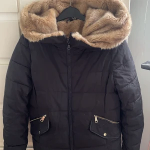Svart pufferjacka  - Svart pufferjacka från Outerwear i storlek S med mjuk beige fuskpäls på insidan och huvan. Jackan har två fickor med dragkedja och guldfärgade detaljer. Perfekt för kalla dagar med sin fluffiga och varma känsla.