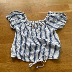 Randig blus från Hollister XS - Blus från Hollister i vitt och blått med ränder. Modellen är croppad med knyt i midjan, korta puffärmar och knappar framtill. 