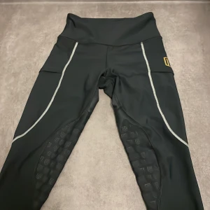 Svarta träningstights från MD - Svarta träningstights från MD med hög midja, gråa kontrastsömmar och guldigt logomärke på låret. Tightsen har greppdetaljer på insidan av benen (som är lite slitna) och är tillverkade i ett stretchigt syntetmaterial som passar perfekt för intensiva träningspass.