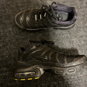Nike Air Max Plus svarta sneakers - Nike Air Max Plus sneakers i helsvart med mesh och syntetpaneler, ikoniska vågiga linjer och synlig Air-dämpning i sulan. Snygga detaljer med gula inslag under sulan och en liten gul etikett med röd Swoosh. Rund tå och snörning för skön passform.