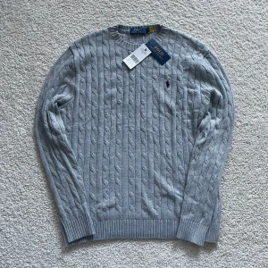 Grå kabelstickad tröja Polo Ralph Lauren - Kabelstickad grå tröja från Polo Ralph Lauren i 100% bomull. Klassisk rund halsringning, ribbade muddar och diskret broderad logga på bröstet. Perfekt för dig som gillar stilren och tidlös design.
