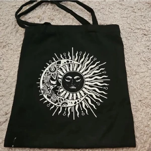 Svart tygkasse med sol och måne-tryck - Cool svart shoppingväska i bomull med ett stort vitt tryck av sol och måne i boho-stil på framsidan. Väskan har långa handtag och är perfekt för att bära böcker, laptop eller shopping. Trycket har detaljer som blommor och mönster som gör den unik. Använd ett par gånger och därför lite smutsig, men inte trasig eller i dåligt skick utöver det. Fick i present för några månader sedan så jag vet tyvärr inte märket.
