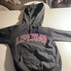 Grå hoodie Lipscomb Champion XS - Mörkgrå hoodie från Champion med stor lila och gul Lipscomb-tryck på bröstet. Klassisk känguruficka, snörning i huvan och mjukt fleecematerial. Perfekt för dig som gillar college-vibe och vill ha något bekvämt och stilrent. Nu pris på denna hooiden ligger på 500kr men jag lägger ut den här för 200kr