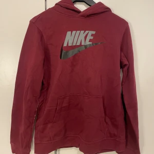 Vinröd Nike hoodie med logga - Säljer en vinröd hoodie från Nike med stor silverfärgad logga på bröstet. Tröjan har huva, magficka och långa ärmar. Perfekt för dig som gillar sportig och avslappnad stil.