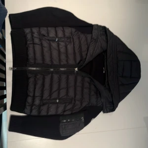 Svart cardigan från Moncler, M - Svart cardigan från Moncler i storlek M. Jackan har stickade ärmar och rygg, quiltad framsida och huva. Dragkedja framtill och fickor med dragkedja på ärmen. Perfekt för kyliga dagar med sin kombination av dun och stickat material är även öppen för byten.