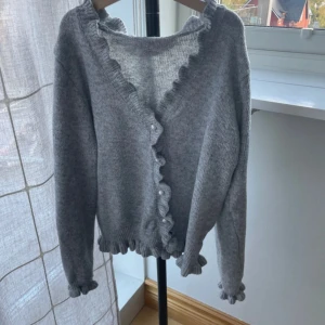 Grå kofta med volang och pärlknappar - Superfin grå kofta med volangkant längs v-ringningen, ärmsluten och nederkanten. Stängs med pärlknappar framtill. Mjuk och stickad i material som känns skönt mot huden. Perfekt för lager-på-lager under kyliga dagar.