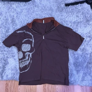 Brun t-shirt med dödskalle och dragkedja - Cool brun t-shirt med korta ärmar, halv dragkedja och kontrasterande krage i rostbrunt. Stor dödskalle-tryck på ena sidan framtill. Skön bomull och perfekt för dig som gillar unika detaljer. Storlek M, bra passform. Aldrig använd.