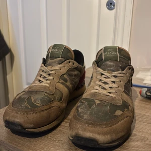 Valentino Rockrunner sneakers camouflage - Säljer ett par Valentino Rockrunner sneakers med ikoniskt camouflagemönster i grönt och beige, röda mockadetaljer på hälen och vita partier bak. Skorna har snörning, platt sula och är tillverkade i en mix av skinn och mocka. Perfekta för dig som gillar streetstyle och designer vibes.