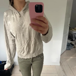 Beige hoodie med stjärnor - Säljer en beige hoodie med huva och snörning. Tröjan har lång ärm och är dekorerad med vita stjärnor på ena axeln. Perfekt för en avslappnad och chill stil.