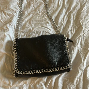 Svart axelväska med kedjedetaljer - Cool svart axelväska i skinnimitation med grov silverkedja runt kanterna och som axelrem. Väskan har en dragkedja på baksidan och ett snyggt, lite rynkat material. Perfekt för dig som gillar edgy detaljer och vill sticka ut.